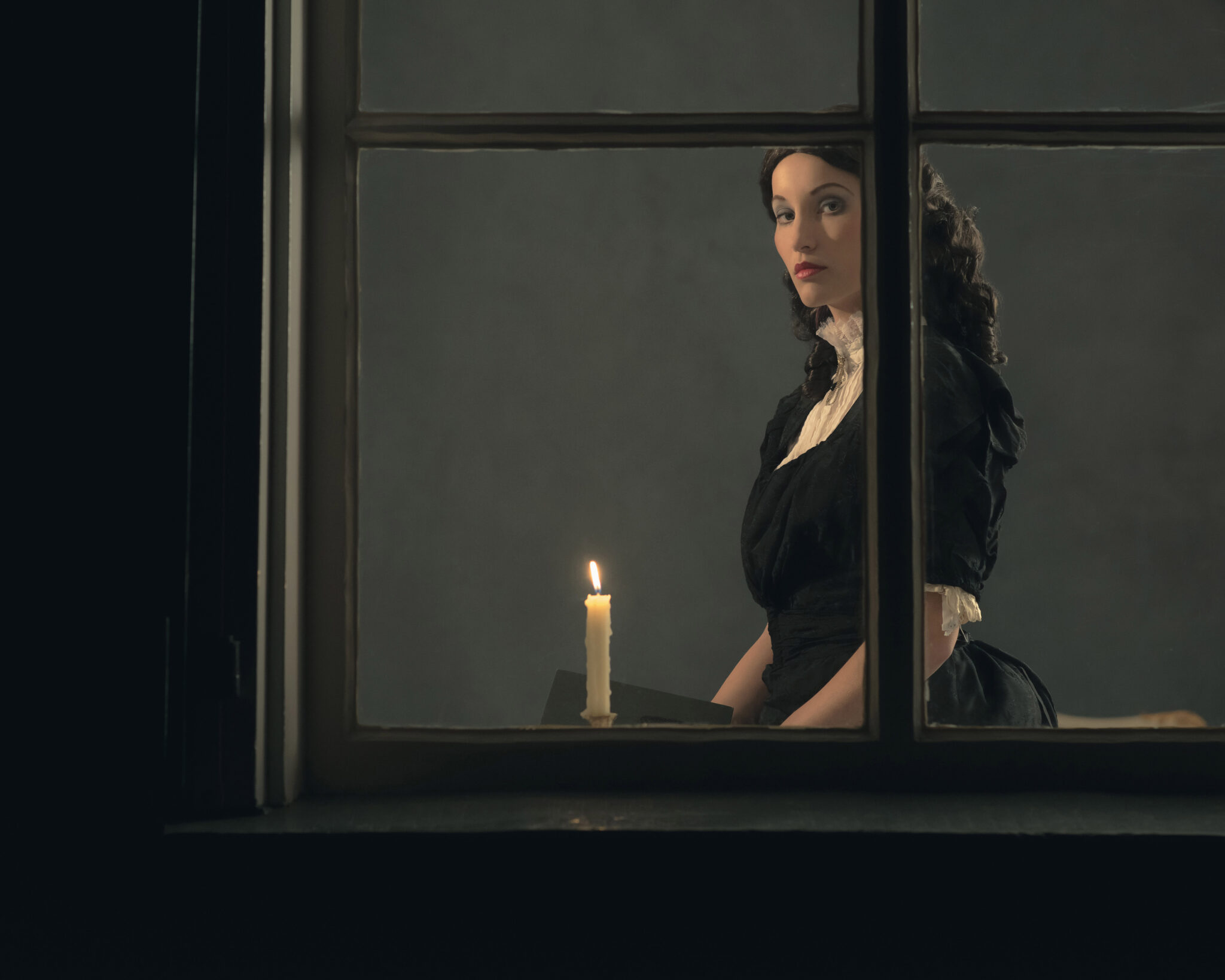 Retro victorian girl reading book by candlelight behind window.